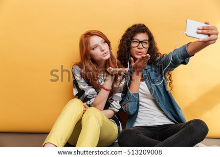 Two cute young women sending kiss and making selfie with mobile phone over yellow background