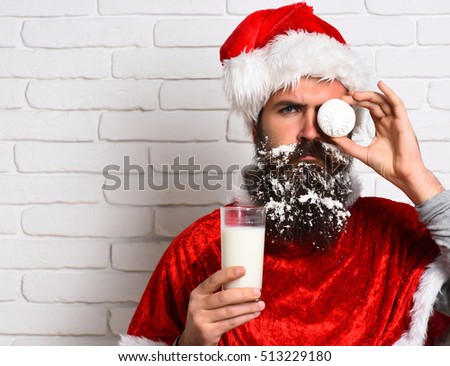 handsome bearded man with stylish mustache and long snowy beard on serious face holding glass of milk with marshmallows instead of eye in red santa suit on white brick wall background, copy space