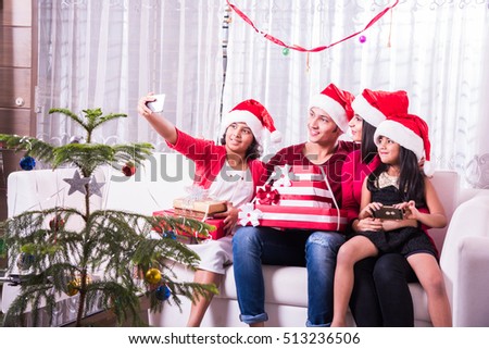 Indian/Asian young family celebrating Christmas with gift while wearing Santa Hat and red Cloths sitting on white couch or sofa. Drinking, taking selfie or decorating Xmas tree. indoor