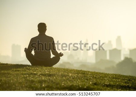 Silhouette of a man sitting in the lotus position meditating on the grassy top of a hill in front of a misty golden sunrise view of the London city skyline