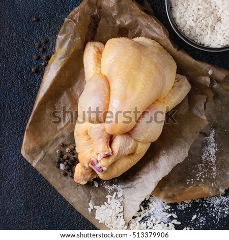 Whole raw mini chicken on baking paper with seasoning and bowl of uncooked white rice over black textural background. Overhead view with space for text. Square image