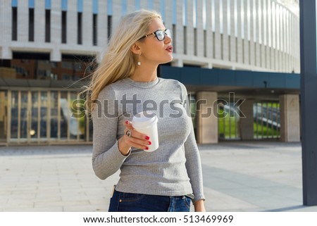 Beautiful attractive young blonde haired businesswoman in glasses, red lips and a white disposable coffee cup, drinking coffee against urban city background. Casual style, shining smile, sunny day. 