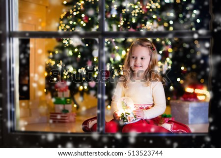 Family on Christmas eve at fireplace. Little girl opening Xmas presents holding snow globe. Child under Christmas tree with gift boxes. Decorated living room. Cozy winter evening at home.