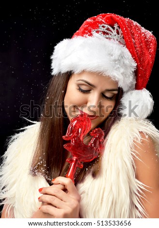 Beautiful woman standing in red christmas santa hat with crown happy smiling and eating sweet sugar cock candy lollipop 2017 symbol on black background