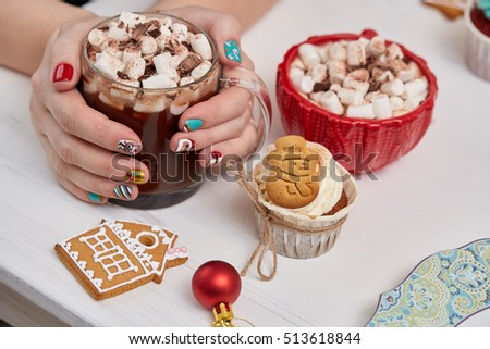 Female hands with bright festive manicure design holding glass mug with hot cocoa and marshmallows. 
Handmade ginger cookies, cupcake,  confection standing on the table. Christmas and New Year treats.