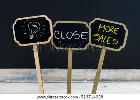 Concept message CLOSE MORE SALES and light bulb as symbol for idea written with chalk on wooden mini blackboard labels, defocused chalkboard and wood table in background