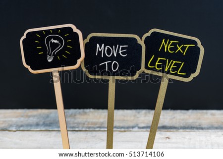 Concept message MOVE TO NEXT LEVEL and light bulb as symbol for idea written with chalk on wooden mini blackboard labels, defocused chalkboard and wood table in background
