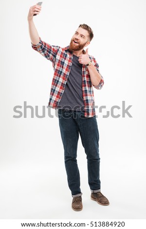 Full length portrait of a smiling casual man taking selfie and showing thumbs up gesture over white background