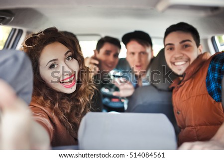 Happy young friends taking photo with a smartphone to her friend sitting back through the window car in a road trip adventure. Female friendship and leisure time concept.