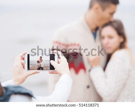 Photographer takes a shot of young couple with a phone on a sea shore in autumn, focus on phone