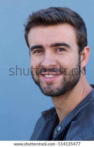 Handsome trendy young bearded man with a friendly smile standing against a blue background looking at the camera, head and shoulders