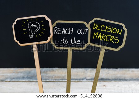 Concept message REACH OUT to the DECISION MAKERS and light bulb as symbol for idea written with chalk on wooden mini blackboard labels, defocused chalkboard and wood table in background