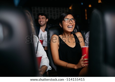 Shot of young woman sitting in multiplex movie theater watching movie and laughing.