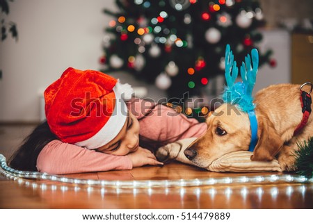 Little Girl with santa hat and and small yellow dog with blue deer antlers laying down by the christmas tree