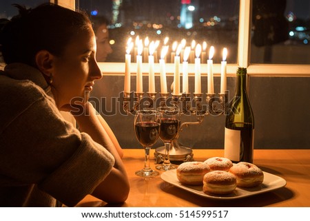 Low key Image of jewish girl celebrating Hanukkah (holiday of lights) with menorah, burning candles, donuts and wine. View to the night Tel Aviv from window.