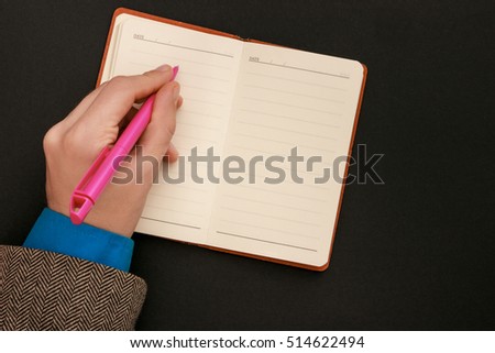 The man writes in a notebook schedule. Pink pen, black background, a man wearing a jacket, pen in hand. Record ideas. Doing Business. left-handed people Royalty-Free Stock Photo #514622494