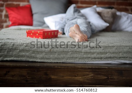 The child lies on the big bed with his face buried in the pillow. Nearby there is a bright red box with a gift. Child isn't happy. He is disappointed with a gift and doesn't want to look at this gift 