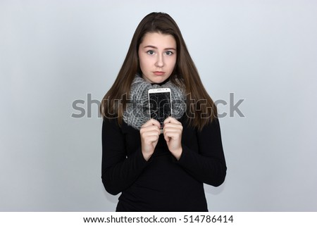 Portrait of a pretty sad woman  in  turtleneck sweaterholding smartphone with broken screen standing over gray background and looking at camera