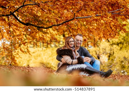 Beautiful smiling couple enjoying in sunny forest in autumn colors. They are sitting on the falls leaves and looking at camera.
