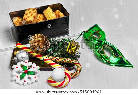 On White wooden background Christmas card of metal box with Christmas cookies, branch of spruce tree, gift, candy cane, Christmas balls, fir-cone, snowflake. Christmas sweets. Christmas carnival mask