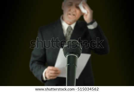 Microphone in sharp focus with a nervous sweaty public speaker or politician blurred in the background preparing to make a speech as he wipes his brow Royalty-Free Stock Photo #515034562