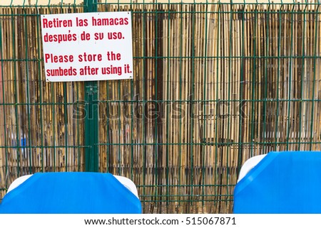 Attention sign with text message in English and Spanish. 
Please store the sunbeds after using it in Spanish language Retiren las hamacas despues de su uso, white sign oustside in garden