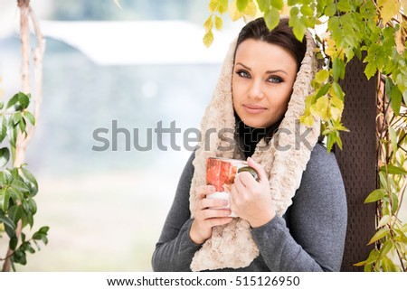 Beautiful green eyed young woman in warm clothes and beige scarf smiling, holding a cup of tea outdoors, in autumn time