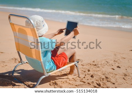 Back view of small cute child playing tablet computer during summer vacation on beach sunny outdoors background