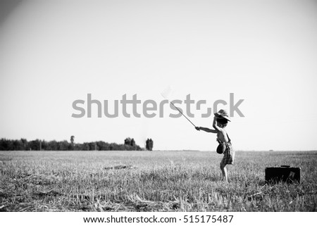 Black and white picture of little boy wearing plaid romper holding insect net on summer countryside background. Side view of kid in safari helmet near old suitcase.