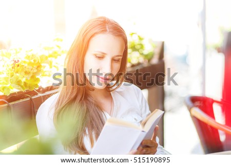 Young woman with long hair reading book at the table in cafe. Pretty caucasian girl with book at the summer terrace