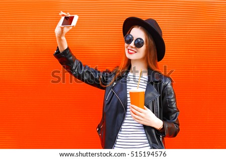 Fashion pretty smiling young woman taking picture self portrait on smartphone in black rock style over city red background
