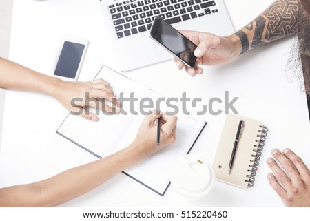 Close up of hipster's tattooed hand holding a smartphone while his colleague is taking notes. 