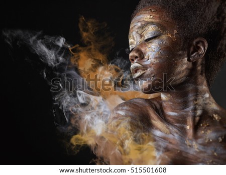 Young African American woman with silver and gold make-up and body art on a black background with smoke Royalty-Free Stock Photo #515501608