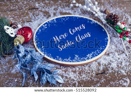 Here Comes Santa Claus Written In Chalk On Blue Chalkboard Holiday Sign Background With Snow And Decorations.