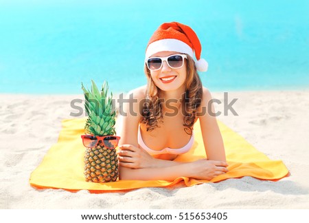 Christmas portrait young smiling woman in red santa hat and pineapple lying on beach over sea background
