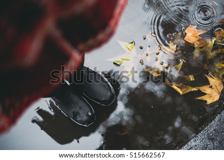 Woman in black rubber boots standing in a puddle with autumn leaves while it's raining. Focus on boots.