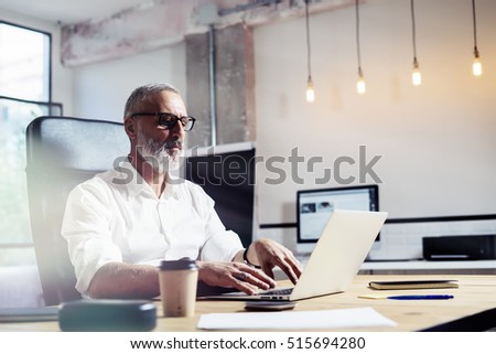 Middle age financial analyst wearing a classic glasses and working at the wood table in modern interior design office.Stylish bearded businessman using laptop on workplace. Horizontal,blurred