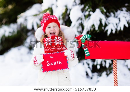Happy child in knitted reindeer hat and scarf holding letter to Santa with Christmas presents wish list at red mail box in snow under Xmas tree in winter forest. Kids sending post to North Pole.