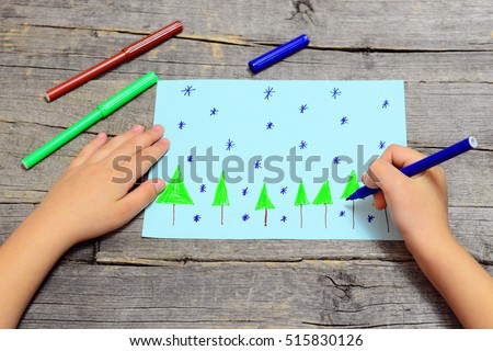 Small child sits and draws Christmas trees and snowflakes on paper. Child holding blue marker in hand and drawing a snowflake. Children drawing, markers on a wooden table. Easy kids art lesson 