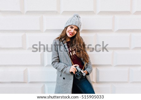 Beautiful young girl with long hair in grey coat and knitted hat on grey wall background. She holds camera in hands and looks enjoyed