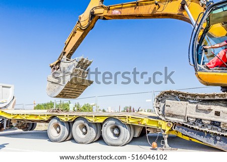 Heavy excavator it climbs on low platform trailer over back ramp, carrying two buckets inserted into one another. Royalty-Free Stock Photo #516046102