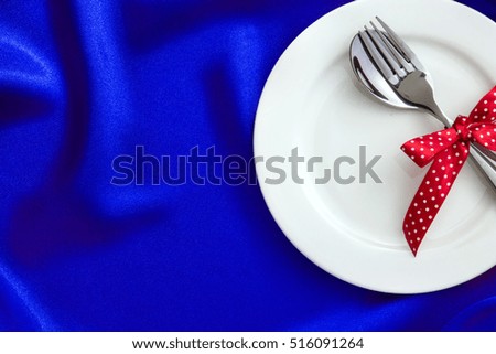  White empty plate with fork and spoon and red ribbon on luxury blue silk fabric for dinner setting 