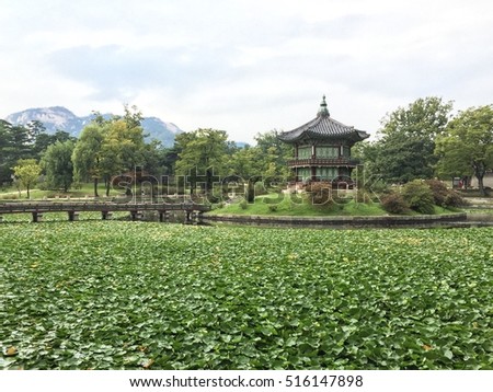 Small Asian shrine standing on a island in a lake full of green plants with a walking bridge over the water with mountains on the background in Seoul, South Korea.