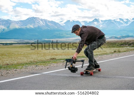 Man rides a longboard and shoot camera with electronic steadycam. Long road in the field on  background of mountains  glaciers.