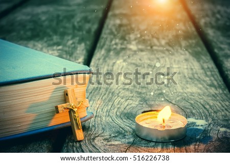 Closeup of wooden Christian cross on bible, burning candle on the old table. Church utensils.