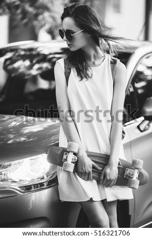 Portrait of lovely urban girl in white dress with a skateboard. Happy smiling woman. Pretty girl is standing near a car. Black and white photography.