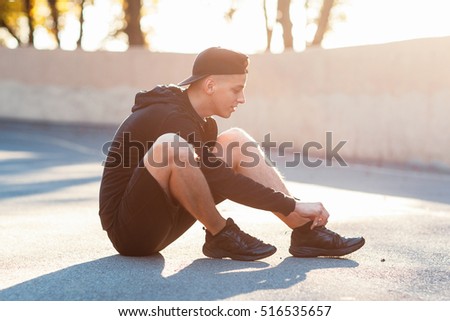 Young sporty man sitting on ground at stadium at sunset. Sportsman preparing for workout before competition, tie shoelaces, blurred photo. Healthy lifestyle, sport, hobby concept