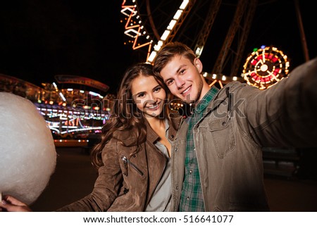 Smiling beautiful young couple with cotton candy taking selfie in amusement park