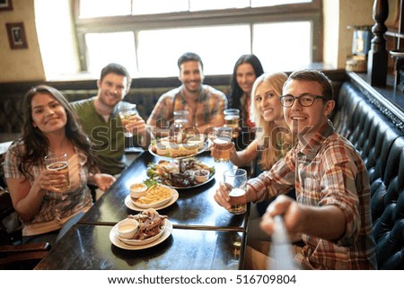 people, leisure, friendship and technology concept - happy friends taking picture by selfie stick, drinking beer and eating snacks at bar or pub