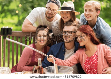 leisure, party, technology, people and holidays concept - happy friends taking picture with smartphone selfie stick and gathering for dinner at summer garden party